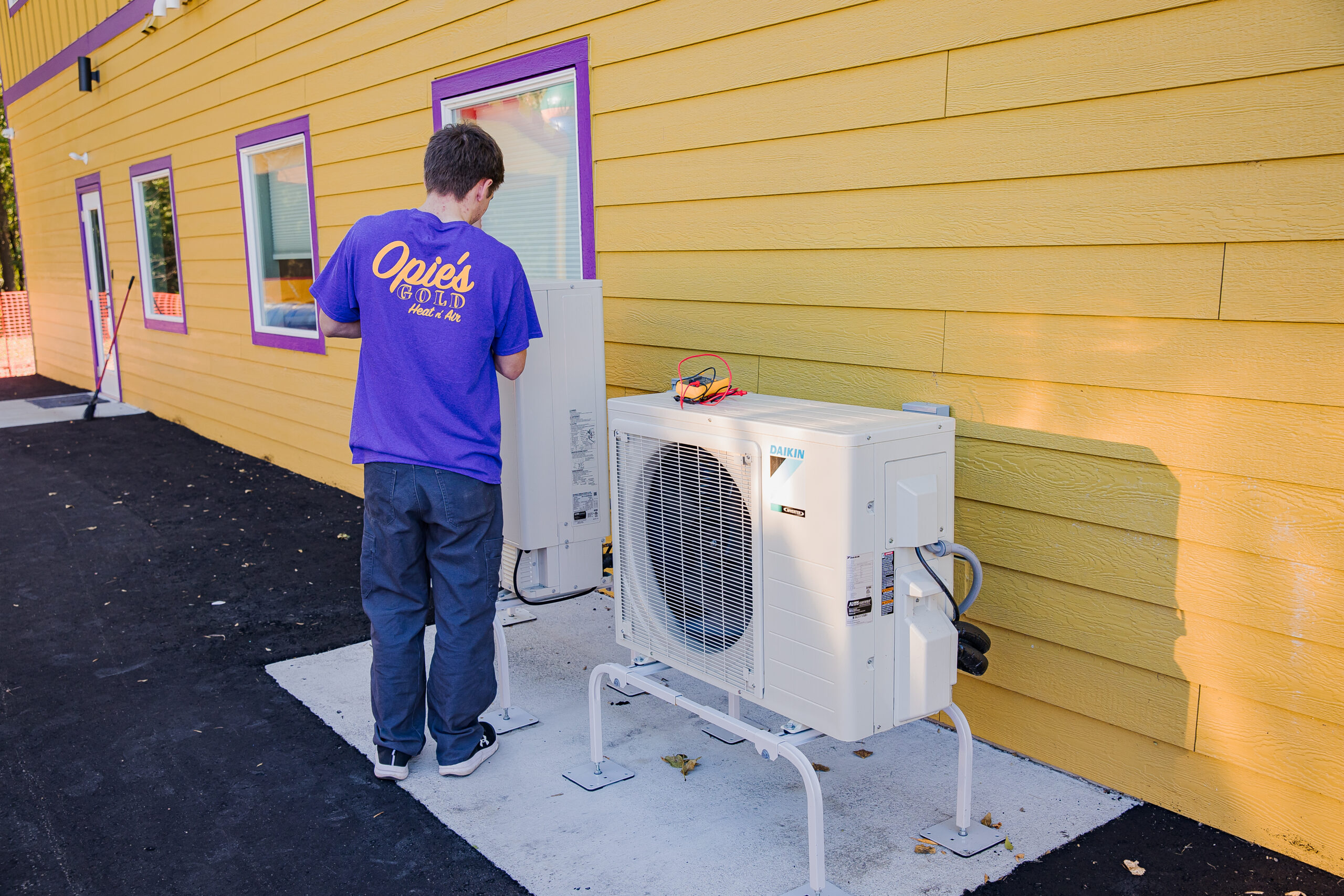 In St. Cloud, a person in a purple shirt diligently repairs an outdoor air conditioning unit attached to a vibrant yellow building.