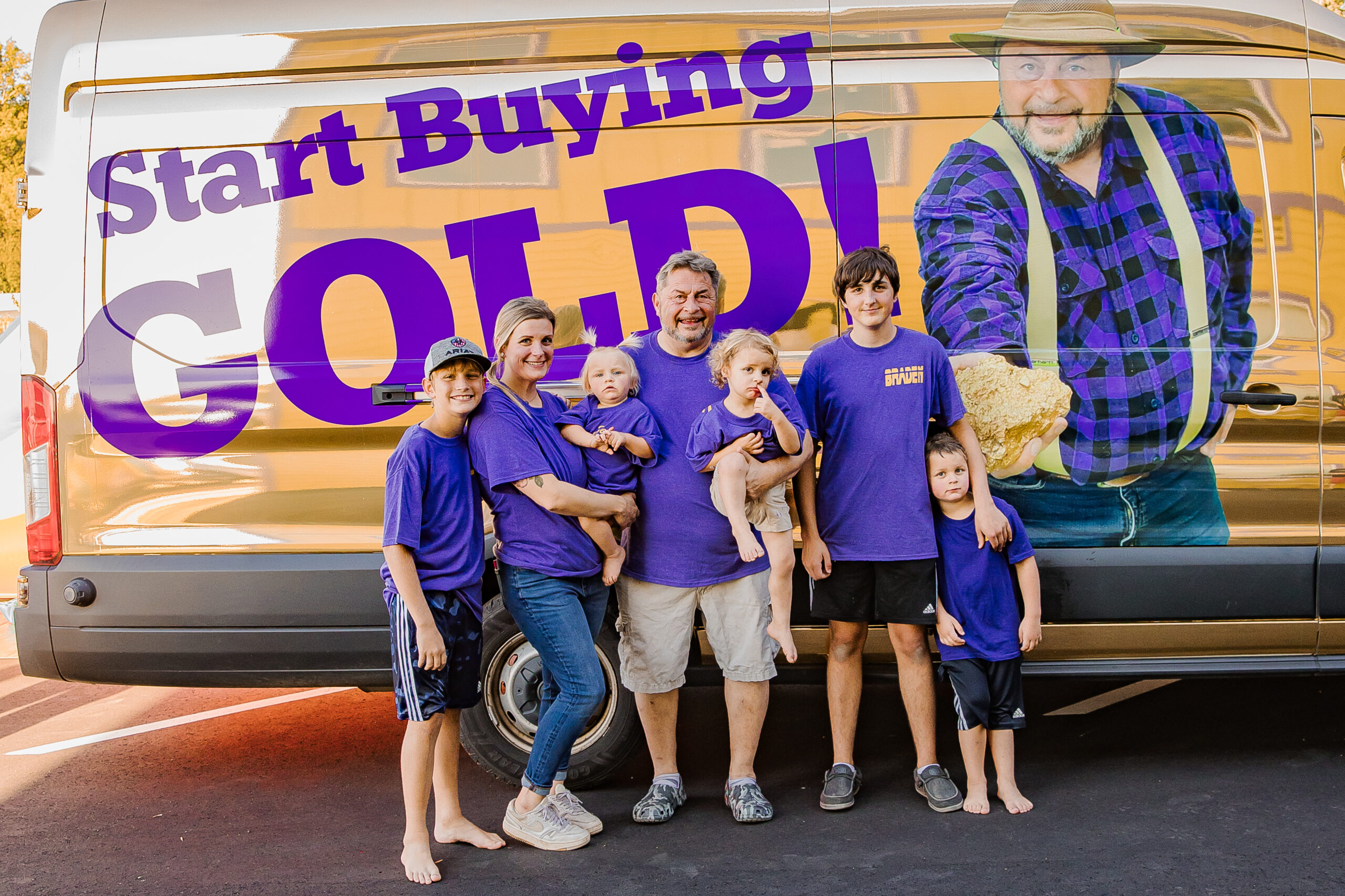 A family of seven, all wearing matching purple shirts, stand in front of a van displaying a large ad with a man holding a gold nugget and the words "Start Buying Gold!