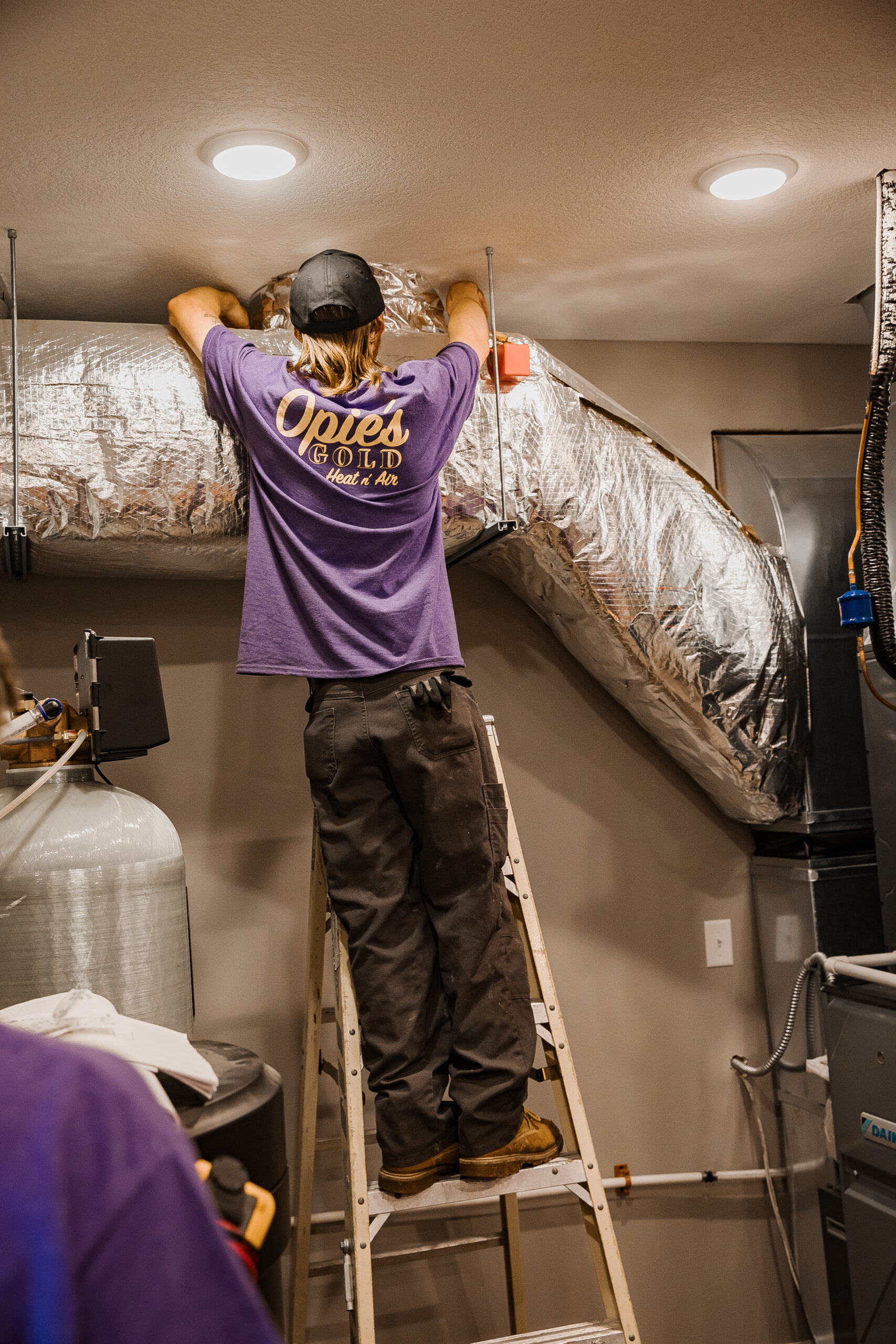 A person in a purple shirt and cap stands on a ladder, adjusting a large silver duct in a room filled with various equipment and pipes.