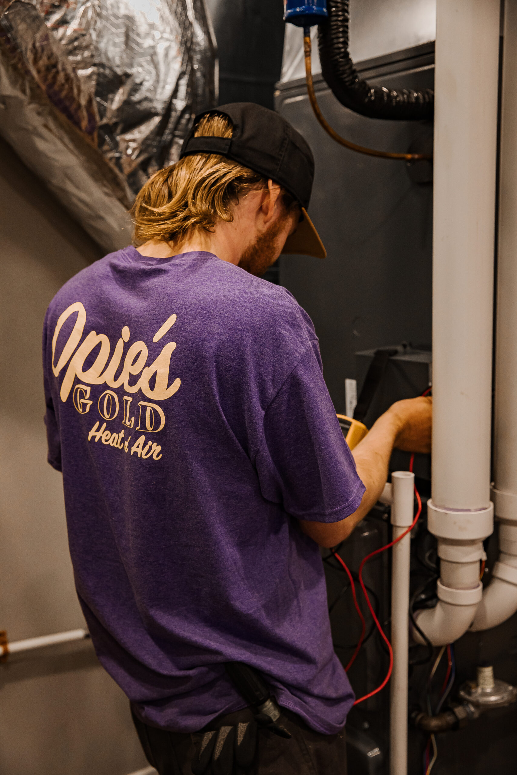 A person in a purple shirt and black cap works on an HVAC system.