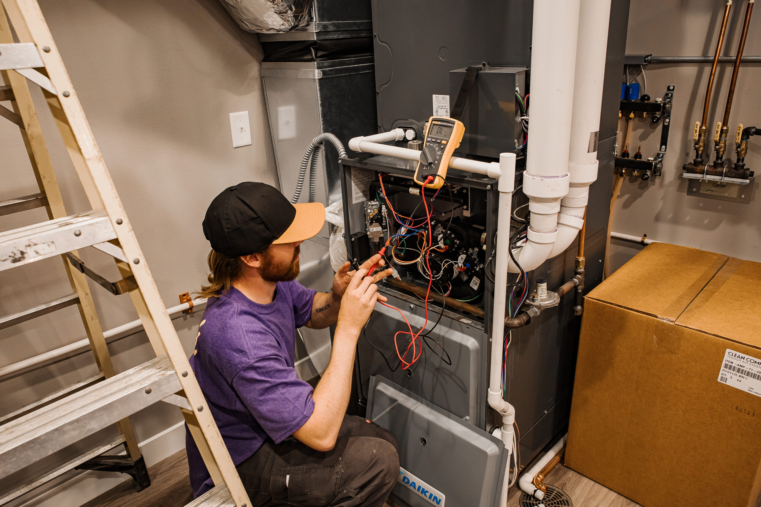 A technician wearing a purple shirt and black cap repairs the furnace using tools and diagnostic equipment, with a ladder and cardboard box nearby.