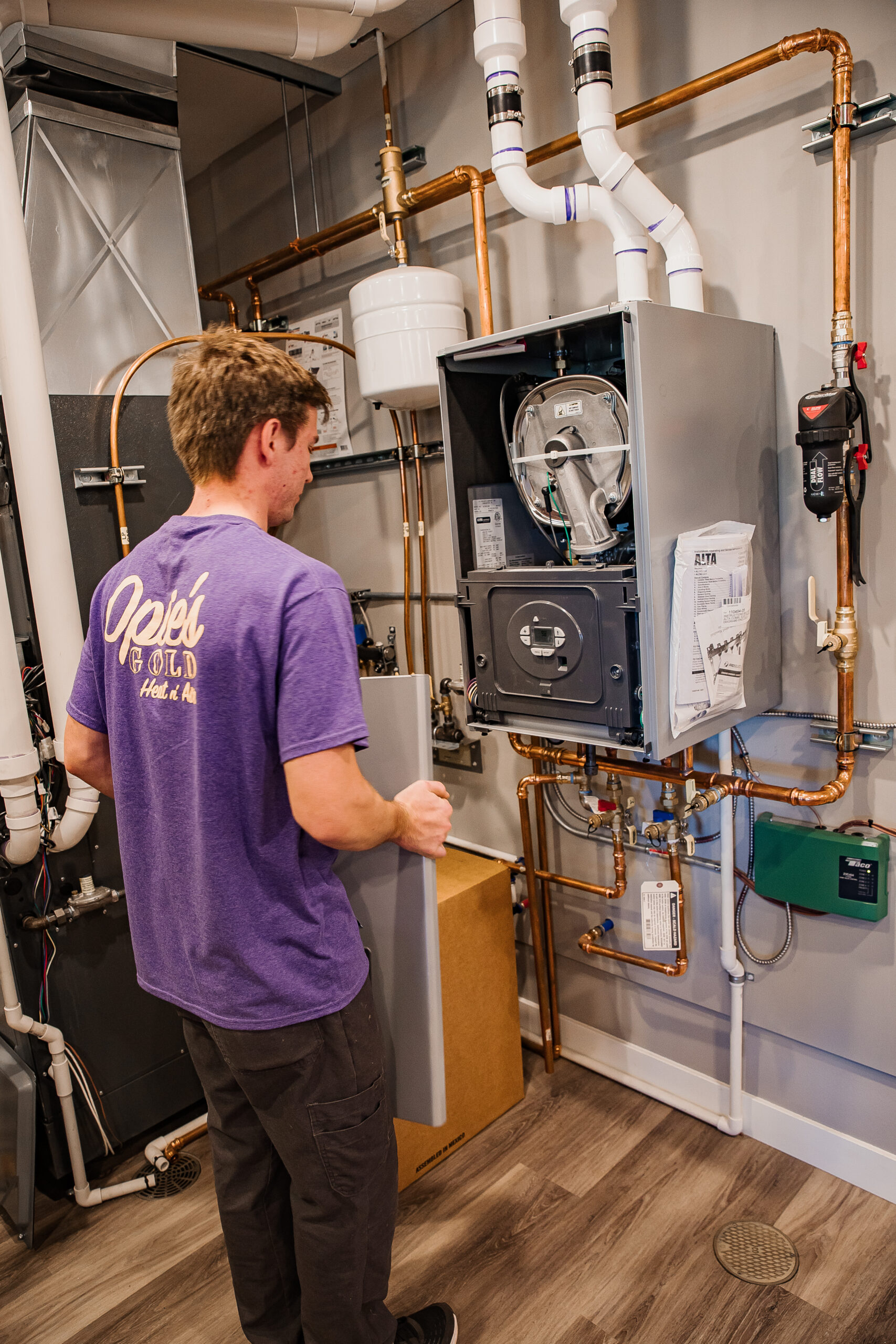 A person in a purple shirt holds a panel and works on a wall-mounted HVAC system. Copper pipes and various components are visible in the setup.