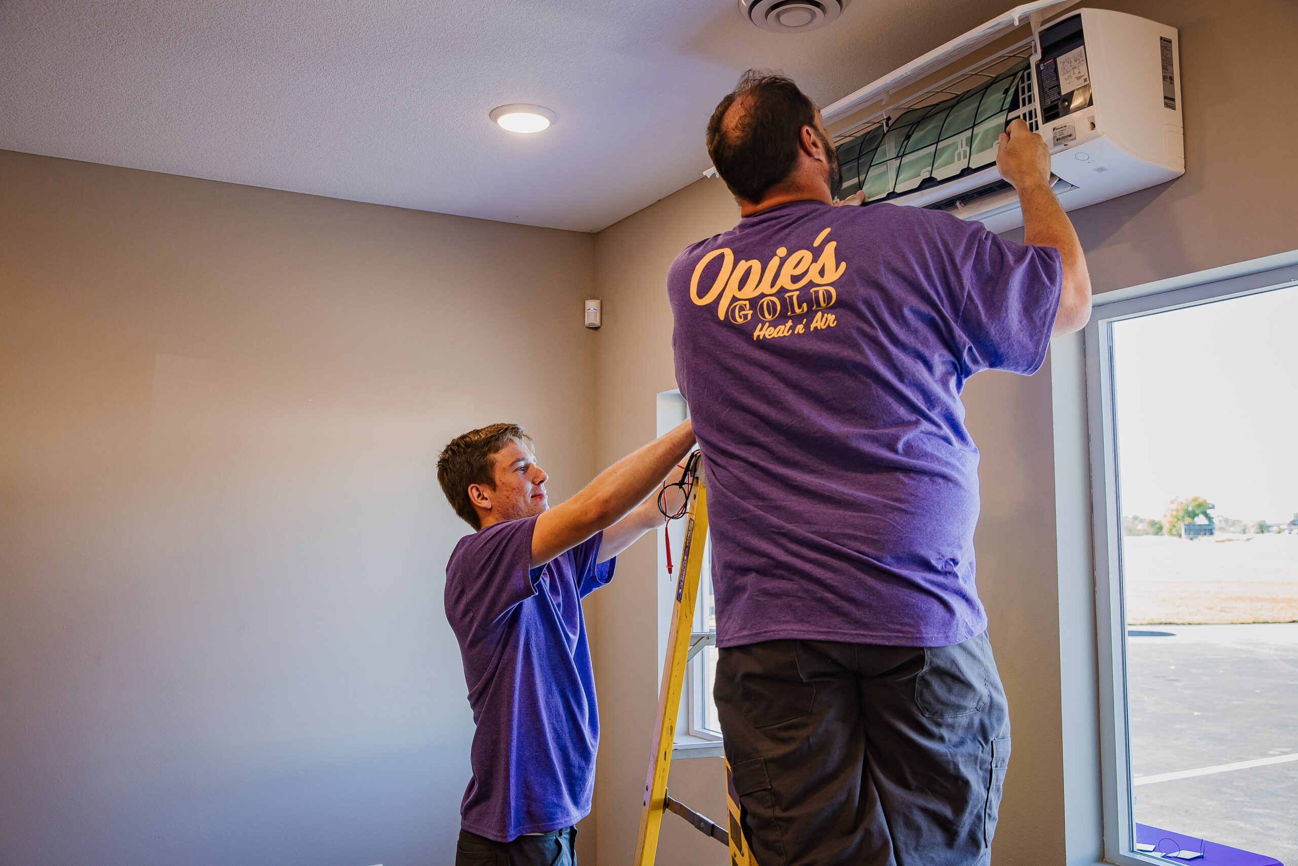 Two technicians in purple shirts install or repair an air conditioning unit inside a room. One climbs a ladder while the other assists.