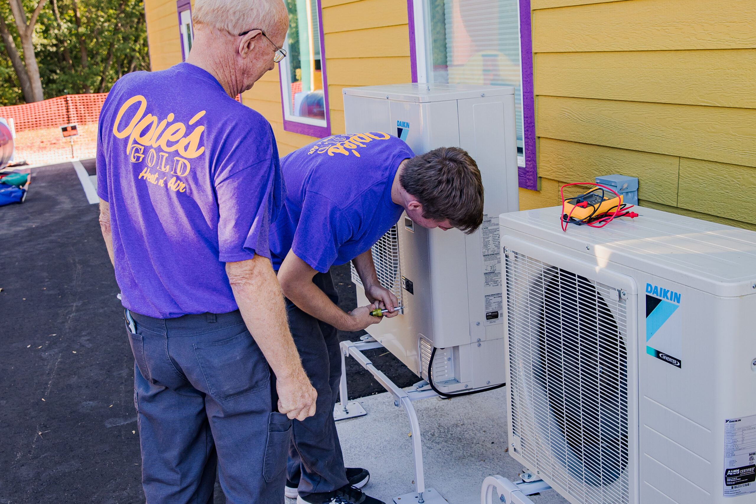 Two men in purple shirts are working on outdoor air conditioning units near a yellow building.