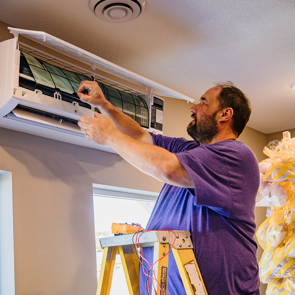 A man stands on a ladder and repairs or inspects an air conditioning unit mounted on an interior wall.