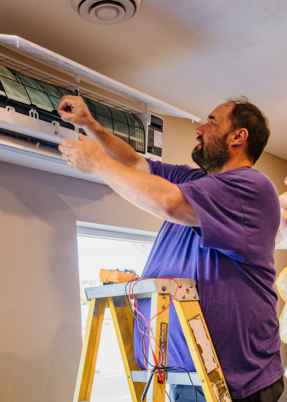 A man stands on a ladder and repairs or inspects an air conditioning unit mounted on the wall indoors.