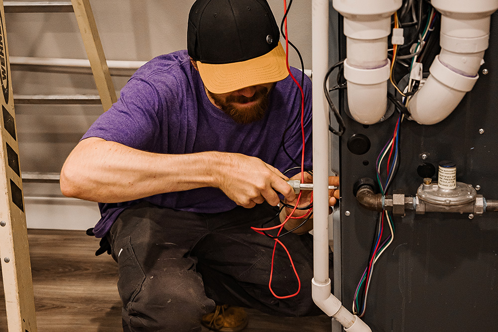 Wearing a purple shirt and cap, a man actively screws wiring inside a mechanical unit with visible pipes and colored wires.