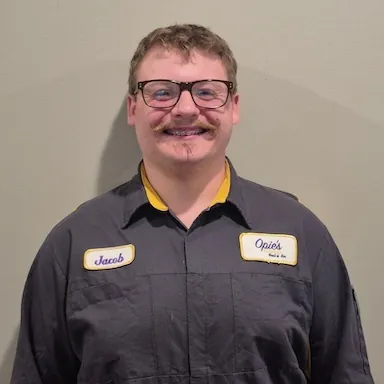 A person in glasses and a dark work uniform with "Jacob" and "Opie's" patches smiles at the camera against a light wall.